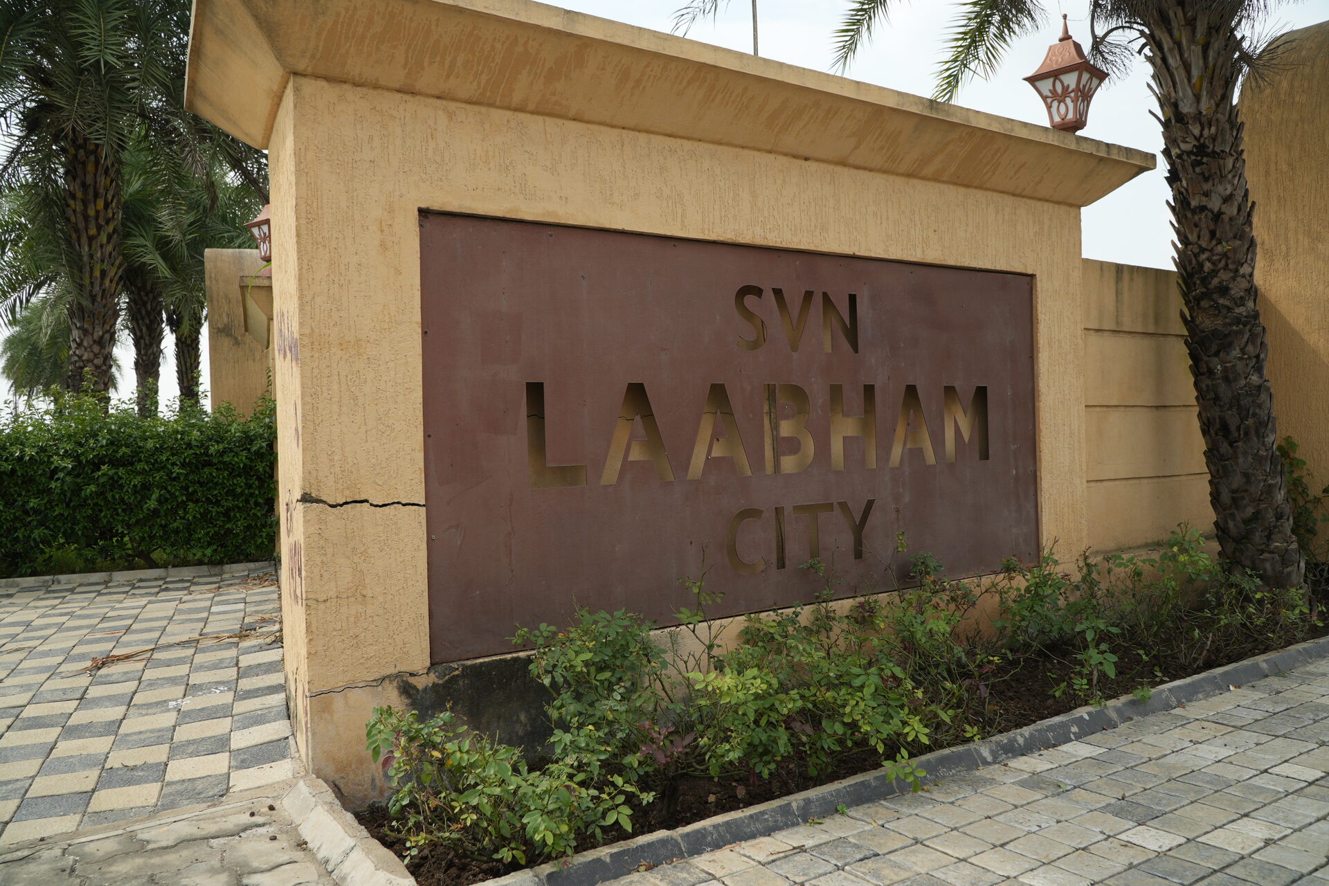 The sandstone entrance wall at Labham City on Super Corridor, the project name cut into a bronze panel, with date palms and a paved footpath beside it.