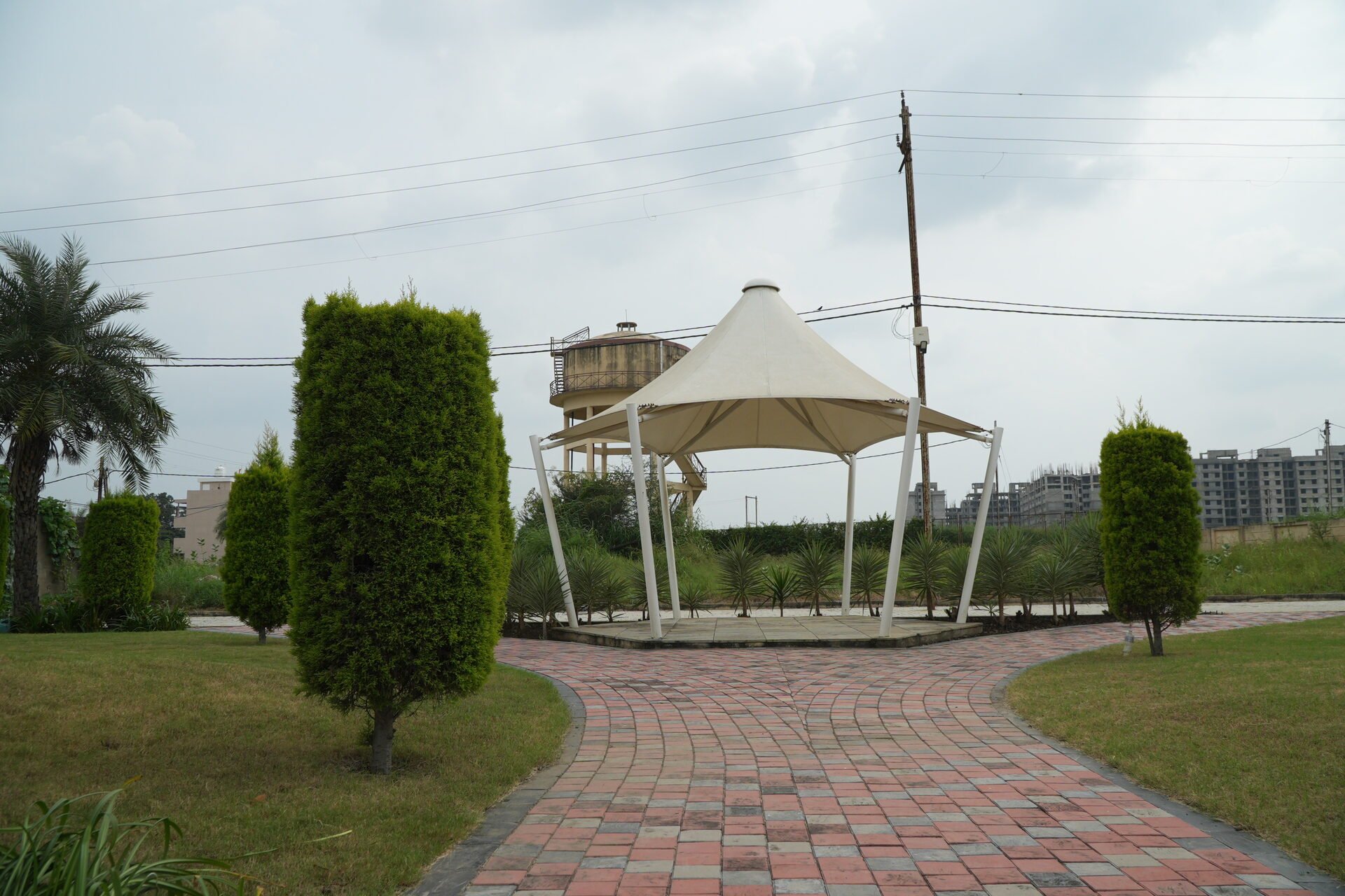 A white canopy pavilion on a paved path at Labham City, among lawns and clipped trees, with a water tank and apartment towers on the Super Corridor skyline.